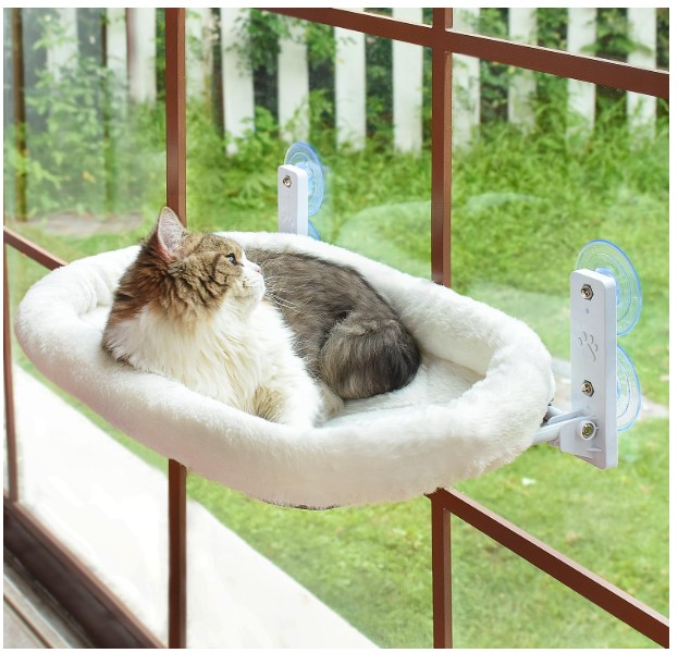 Fluffy tabby cat lounging in a white fleece-lined window perch hammock with suction cup mounts overlooking a green backyard