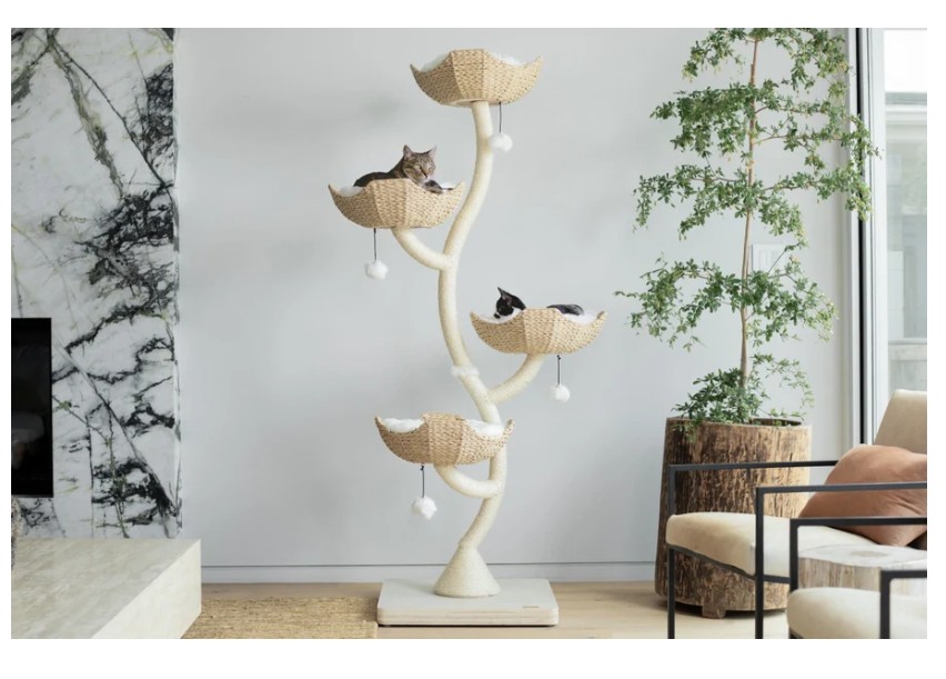 Two cats lounging in woven basket-style perches on a sculptural cat tree with natural textures in a modern living room with marble and greenery