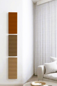 Three rectangular wall-mounted cardboard cat scratchers in warm earth tones displayed vertically on a white wall in a minimalist living room with a modern sofa and sheer curtains.